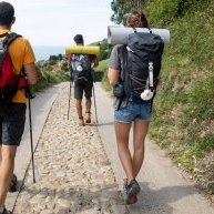 três jovens fazem o percurso do Caminho de Santiago com mochilas às costas numa rua rural empedrada